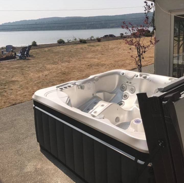Outdoor hot tub on a concrete patio overlooking a serene lake, with lounge chairs and a fire pit in the distance under a hazy sky.