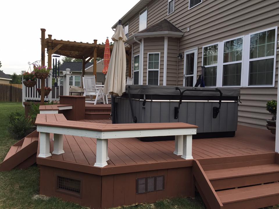 Spacious wooden deck attached to a suburban home, featuring a covered hot tub, built-in seating, a pergola with patio furniture, and hanging flower baskets.