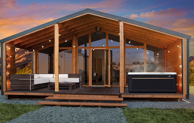 Modern wooden cabin with a covered deck, featuring string lights, outdoor seating, and a steaming hot tub, set against a scenic sunset backdrop.