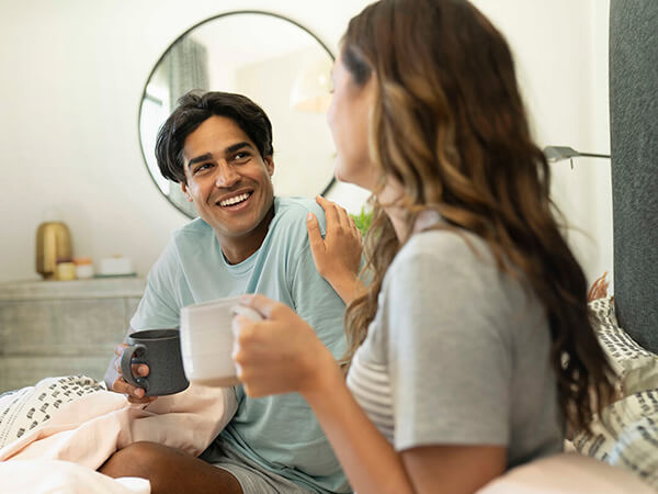 Smiling couple enjoying a conversation while drinking coffee in bed, with a cozy and stylish bedroom setting in the background.