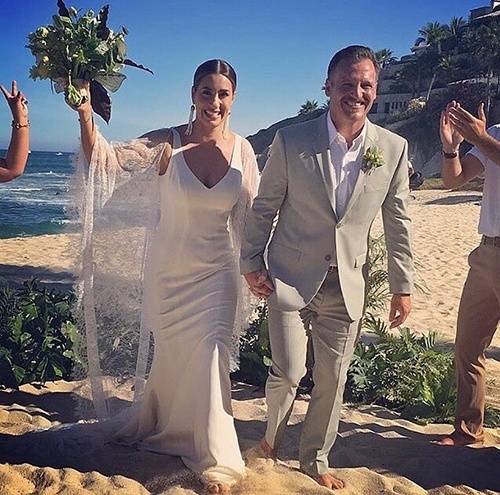 a newlywed couple in formal attire walk barefoot across a beach celebrating their new marriage