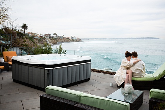 A couple relaxes on an ocean side patio with their perfect hot tub in the background