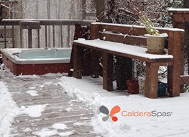 a hot tub sits on a deck in a snowfall surrounded by a snowy forest
